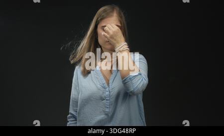 Young blonde woman pinches bridge of nose and closes eyes under harsh lighting in black studio; disgust. Stock Photo