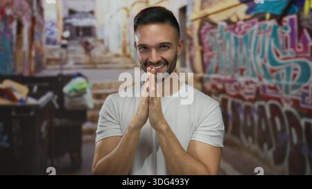 Young hispanic man standing outdoors in a graffiti-covered urban street with a thoughtful expression, wearing a white t-shirt, surrounded by vibrant c Stock Photo