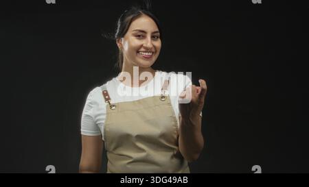 Woman in apron smiles and beckons with right hand in studio set with black backdrop; friendly invitation. Stock Photo