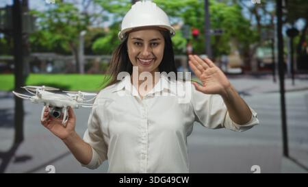 Woman engineer holding drone and waving hand on street wearing hardhat; confidence innovation teamwork. Stock Photo
