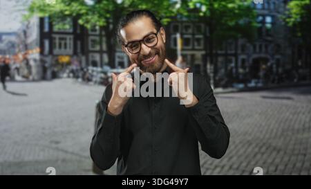 Man wearing glasses and black shirt points index fingers to cheeks and smiles showing teeth on a cobbled city street; joy easy charm. Stock Photo