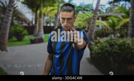 Man in blue and black soccer jersey pointing a forefinger at camera on a palm lined resort path; defiance challenge. Stock Photo