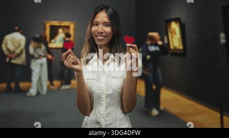 Young thai woman smiles and holds two small red felt hearts between her fingers while standing in a public building gallery with visitors viewing fram Stock Photo