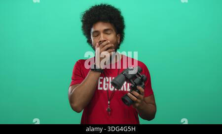 Tired lifeguard man holding binoculars against vivid green background yawns, blending roles of vigilance and fatigue. Stock Photo