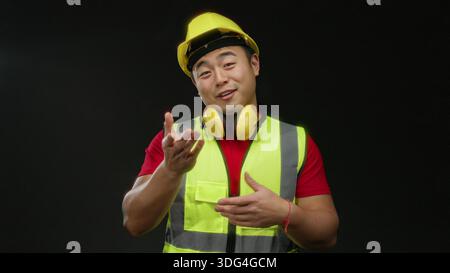 Man wearing safety vest and hard hat gestures playfully against a black background, showcasing confidence and safety awareness on site. Stock Photo