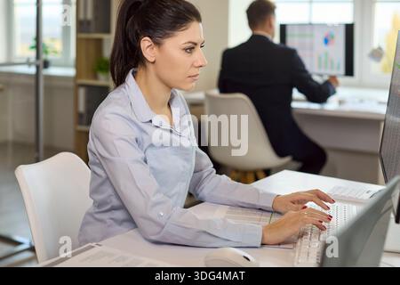 Focused office analyst typing financial data on computer while colleague works Stock Photo