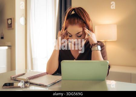Young woman experiencing mental fatigue while working on a digital tablet and notebook indoors, showing stress, concentration problems, cognitive over Stock Photo