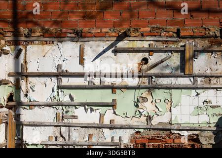 A deteriorated building wall featuring exposed red bricks, peeling plaster, remnants of old paint, and wood framework. Stock Photo