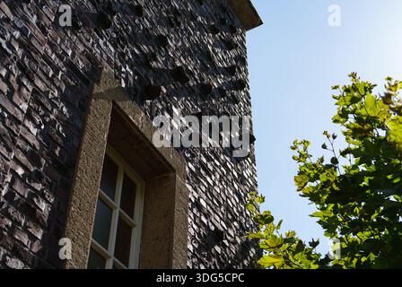 Angled view of a textured stone wall with a window, framed by bright green leaves against a clear sky Stock Photo