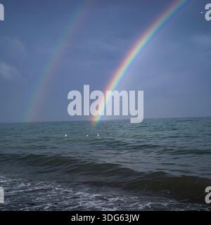 A vivid rainbow arcs over a calm ocean with gentle waves under a gray sky. Qualicum Beach, British Columbia, Canada Stock Photo
