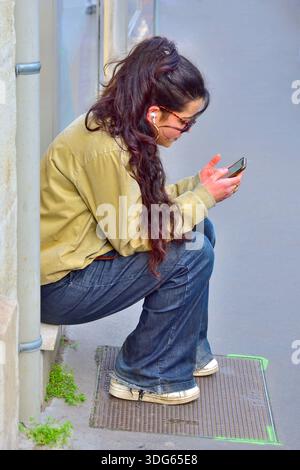 Young woman / teenager / student sitting on stone step whilst messaging on mobile phone - Tours, Indre-et-Loire (37), France. Stock Photo