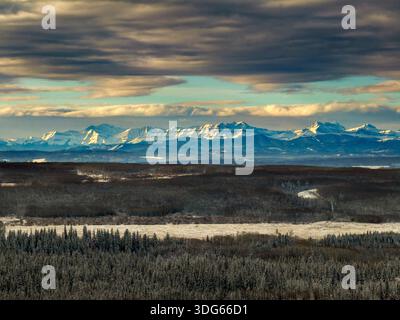 Snow-capped mountains under dramatic clouds with a forest and plains in the foreground. Calgary, Canada Stock Photo