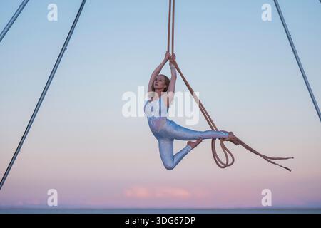 Aerial dancer in a striking pose, suspended by ropes during sunset over calm sea. WA, USA Stock Photo