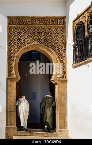 Two people in traditional attire enter an ornate arched doorway with intricate carvings. Rabat, Morocco Stock Photo