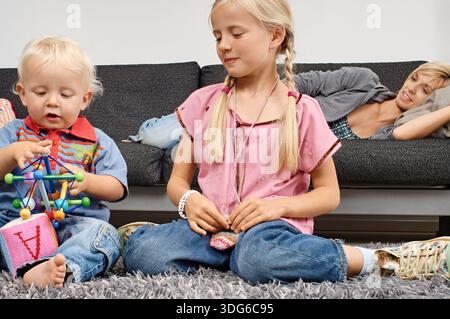 Two children playing on a rug with a woman relaxing on the couch in the background. Germany Stock Photo