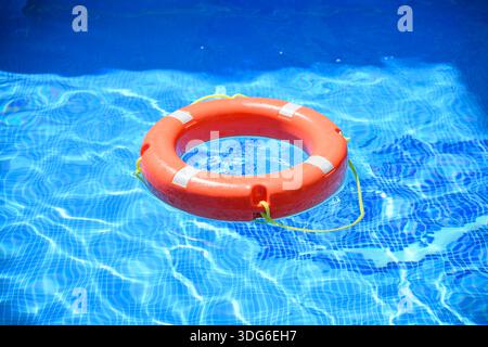 Bright orange life preserver floats in pool water, sun's rays creating shimmering waves. Lifesaving equipment ensures safety. Drowning prevention. Wat Stock Photo