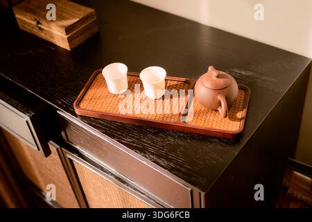 Wooden tray with brown teapot and two white cups on dark cabinet surface, warm lighting. Trip from Guilin to Chengdu, Guangxi, Sichuan, China Stock Photo