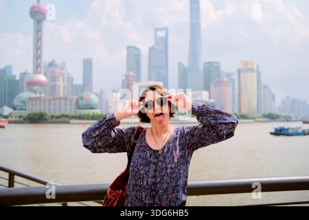 Woman posing playfully with sunglasses in front of a city skyline and river. Shanghai, China Stock Photo