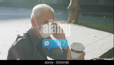 Talking mature man on smartphone holding to-go cup, wearing dark shirt white N95 mask sunlit plaza Stock Photo