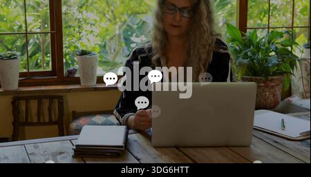 Working mature woman wearing dark top and eyeglasses typing on laptop in sunroom, notebooks beside Stock Photo