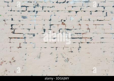 Brick wall painted in light beige with uneven coverage and signs of wear. Dark stains and remnants of old paint emphasize age and deterioration. Stock Photo