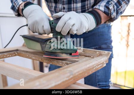 Craftsman. Adult carpenter using an electric sander to smooth an old wooden window. Construction industry, carpentry, housework do it yourself, workpl Stock Photo