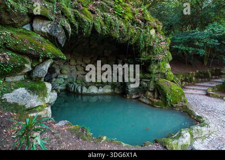 A tranquil, turquoise pool is nestled within a grotto of moss-covered rocks and stone walls, with a cobblestone path leading to steps in a serene, for Stock Photo