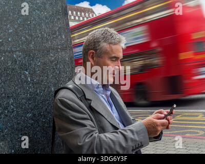 UBER TAXI APP TEXT Mature businessman 55-60 yrs using smartphone central Southbank  London red bus with speed blur passing in background London UK TFL Stock Photo