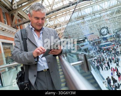iPad Businessman using smart tablet on busy commuter commuting rail Waterloo Station railway concourse with clock, business on the move.  London UK Stock Photo
