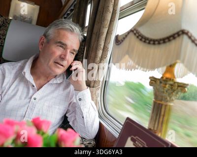 LUXURY TRAVEL LIFESTYLE TRAIN FIRST CLASS  Relaxed man 55-60yrs, talking on smartphone enjoying trip in luxury Pullman train dining rail carriage Stock Photo