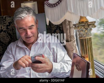 LUXURY TRAVEL LIFESTYLE TRAIN FIRST CLASS Relaxed man 55 yrs texting smartphone enjoying luxury Pullman train dining menu table rail carriage at speed Stock Photo