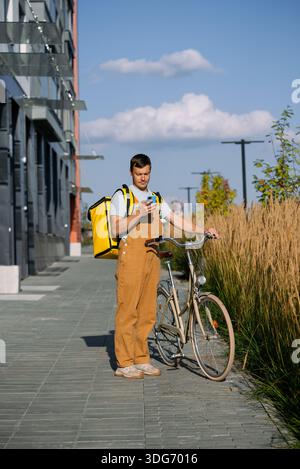 Young male bike courier in brown overalls and yellow delivery bag stands with bicycle on city street, checking smartphone app for food orders and navigation during daylight Stock Photo