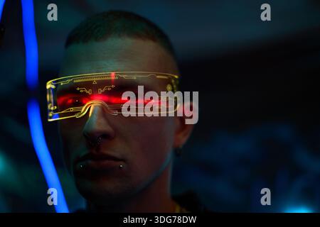 Portrait of young adult Caucasian man wearing futuristic augmented reality glasses with glowing yellow interface, standing in dark environment with neon blue and red lights reflecting on face Stock Photo