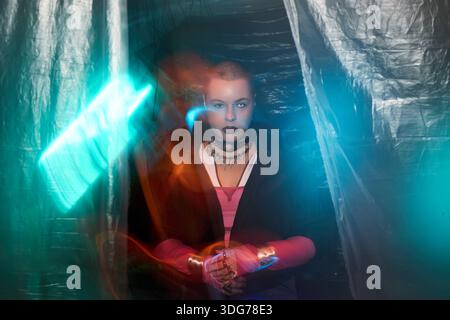 Portrait of young Caucasian woman sitting in dimly lit room with intense gaze, holding hands together, surrounded by digital neon light effects and plastic sheets, embodying cyberpunk hacker concept Stock Photo