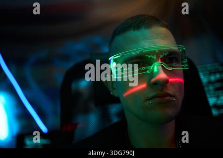 Portrait of young adult Caucasian man, wearing futuristic augmented reality transparent visor glasses, sitting in front of computer monitors with digital code reflecting on face in dark room Stock Photo