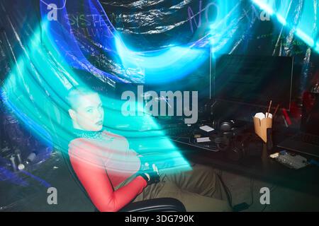Young adult Caucasian woman with shaved head sitting at desk using computer surrounded by monitors and electronic equipment, neon blue light trails illuminating hacker workspace Stock Photo