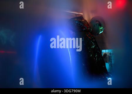 Young adult Caucasian man wearing reflective jacket and neon glasses standing in dark room using computer equipment, illuminated by blue and red lights, engaging in hacking activity Stock Photo