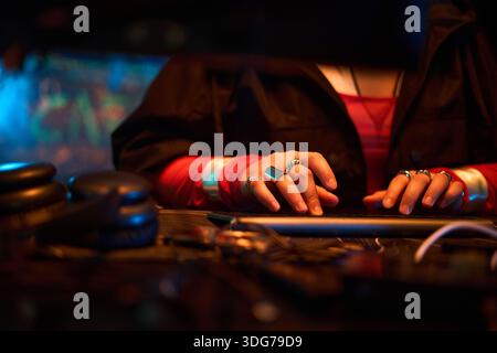 Young adult Caucasian woman typing on computer keyboard with hands visible, wearing multiple rings, engaging in hacking activity in dark room with electronic equipment visible Stock Photo