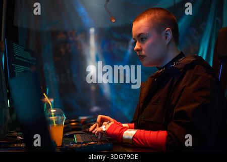Young adult Caucasian woman with shaved head and facial piercings sitting at desk typing on keyboard, concentrating on computer screens with code in dark room, plastic cup nearby Stock Photo