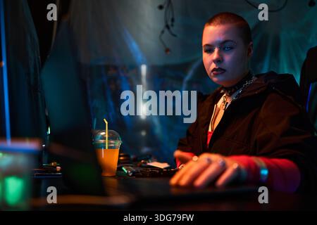 Portrait of young adult Caucasian woman with shaved head sitting at desk using laptop, hand on keyboard, focused expression, illuminated by blue and orange light, plastic sheet background Stock Photo