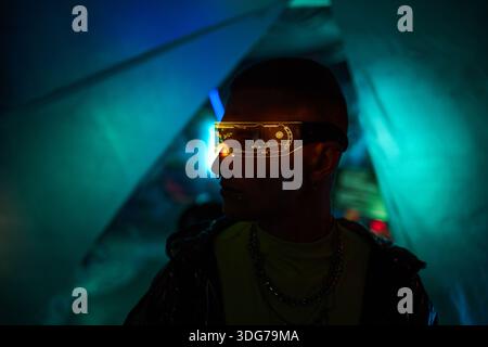 Young adult Caucasian man wearing futuristic illuminated visor standing in dark room with neon blue and green lighting, facing sideways, displaying hacker and cyber club concept Stock Photo