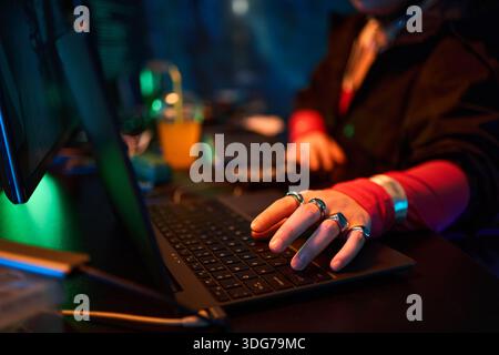 Hand of anonymous young woman typing on laptop with multiple rings on fingers, sitting at desk with another computer and drink in background, engaging in hacking activity Stock Photo