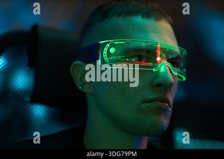 Caucasian young adult man wearing futuristic augmented reality glasses sitting indoors, displaying digital interface reflections on lenses, showing focused facial expression Stock Photo