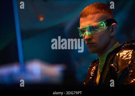 Young adult Caucasian man wearing futuristic augmented reality glasses sitting at computer workstation hacking digital system in dark room with neon lighting Stock Photo