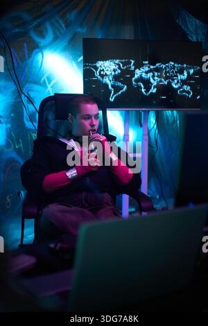 Caucasian young adult man sitting in dark room drinking from bottle while looking at computer screen with digital world map, wearing hoodie and fingerless gloves, illuminated by blue light Stock Photo
