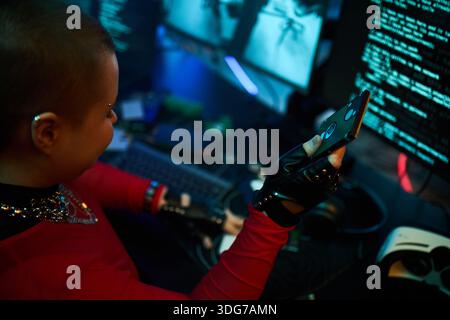 Young adult Caucasian woman sitting at computer using smartphone or holding hard drive while typing on keyboard with gloved hand, multiple monitors displaying code in dark room, engaging in hacking Stock Photo