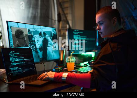 Young adult Caucasian woman with short hair sitting at desk working on multiple computers, monitoring surveillance footage and writing code, focused on cybersecurity hacking activity Stock Photo