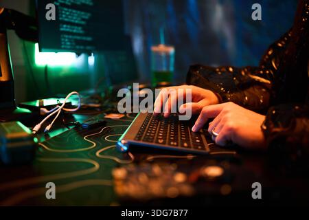 Hands of young man typing on computer keyboard with programming code on monitor, sitting at desk in dark room, hands visible, engaging in hacking activity, electronic devices nearby Stock Photo