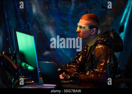 Young adult Caucasian man wearing futuristic glasses sitting at workstation using multiple computer monitors, concentrating on screen in dark room with cyber elements visible Stock Photo