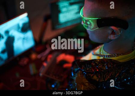Young adult Caucasian man wearing futuristic glasses sitting at desk typing on keyboard with multiple monitors displaying code and digital information in dark room Stock Photo